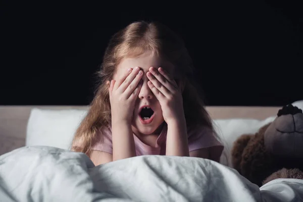 Enfant effrayé crier et couvrir les yeux avec les mains tout en étant assis sur la literie près ours en peluche isolé sur noir — Photo de stock