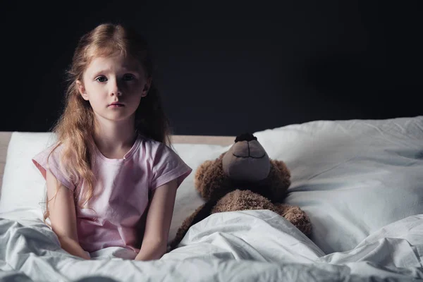 Criança assustada sentado na cama perto de ursinho de pelúcia e olhando para a câmera isolada no preto — Fotografia de Stock