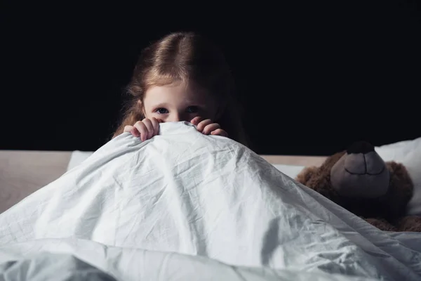 Scared child hiding under blanket while sitting on bedding near teddy bear isolated on black — Stock Photo