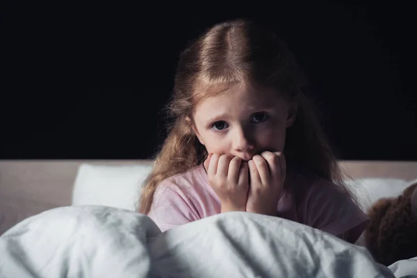 Bambino spaventato guardando la fotocamera mentre seduto su biancheria da letto isolato su nero — Foto stock