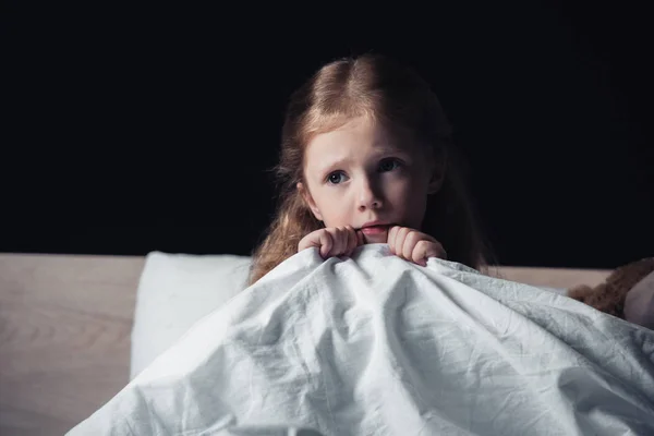 Scared child looking away while sitting on bedding in dark bedroom isolated on black — Stock Photo