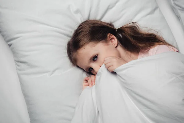 Top view of scared child lying on white bedding and looking at camera — Stock Photo