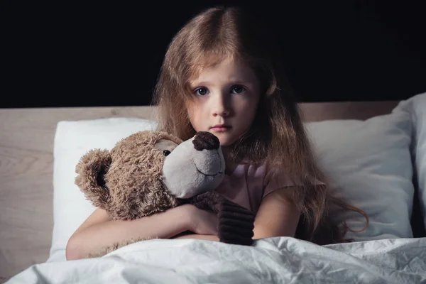 Испуганный ребенок обнимает плюшевого мишку, сидя под одеялом и глядя на камеру, изолированную на черном — стоковое фото