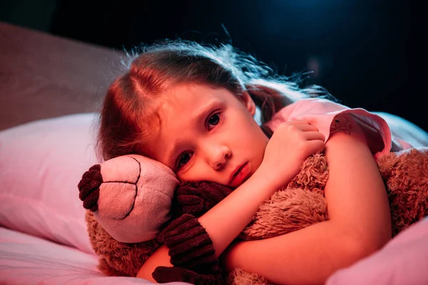 Испуганный ребенок обнимает плюшевого мишку, лежа в постели и глядя на камеру, изолированную на черном — стоковое фото
