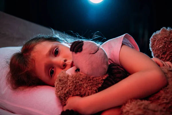 Перелякана дитина обіймає плюшевого ведмедя, лежачи в ліжку і дивлячись на камеру — стокове фото