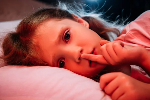 Испуганный ребенок лежит в темной спальне, показывает знак молчания и смотрит в камеру — стоковое фото