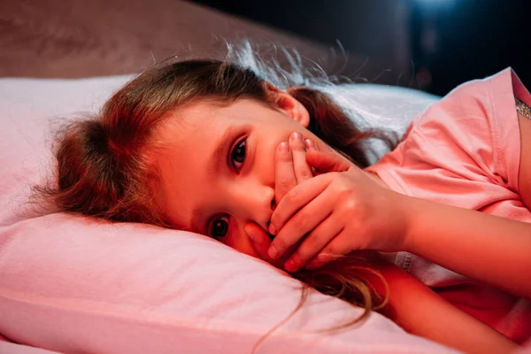 Scared child looking at camera while lying in bed and showing hush sign — Stock Photo