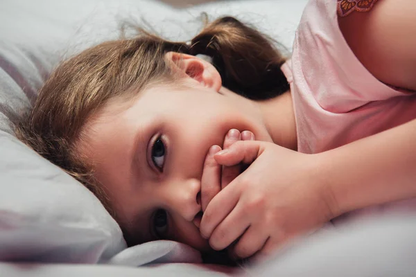 Bambino spaventato guardando la fotocamera mentre sdraiato a letto e coprendo la bocca con le mani — Foto stock