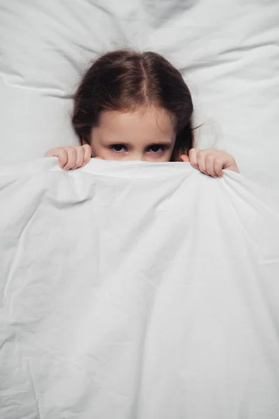 Вид испуганного ребенка, прячущегося под одеялом и смотрящего в камеру — стоковое фото