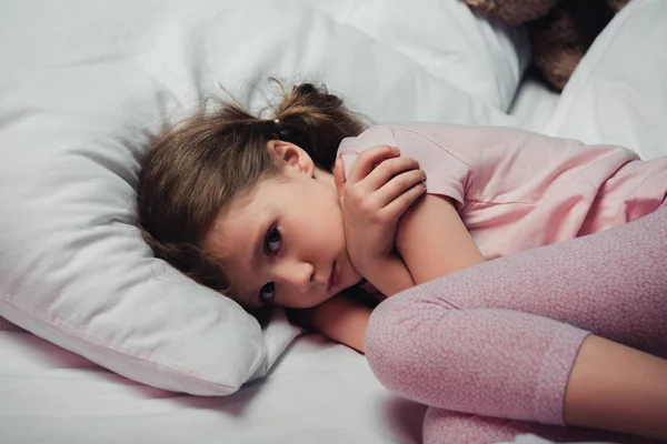 Niño asustado mirando a la cámara mientras está acostado en la cama y abrazándose con los brazos - foto de stock