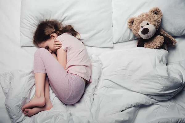 Вид зверху на налякану дитину, що лежить на білому постілі біля плюшевого ведмедя — стокове фото