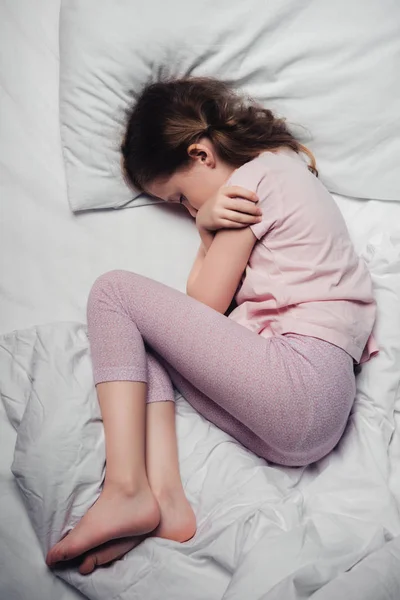 Top view of scared child lying in bed and hugging herself with arms — Stock Photo