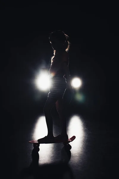 Kid riding penny board in darkness with illumination of headlights on black background — Stock Photo