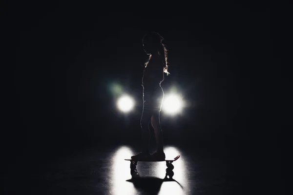 Child riding penny board in darkness with illumination of headlights on black background — Stock Photo