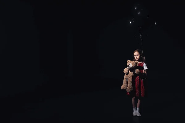 Deprimido, criança solitária segurando balões pretos e ursinho isolado em preto — Fotografia de Stock
