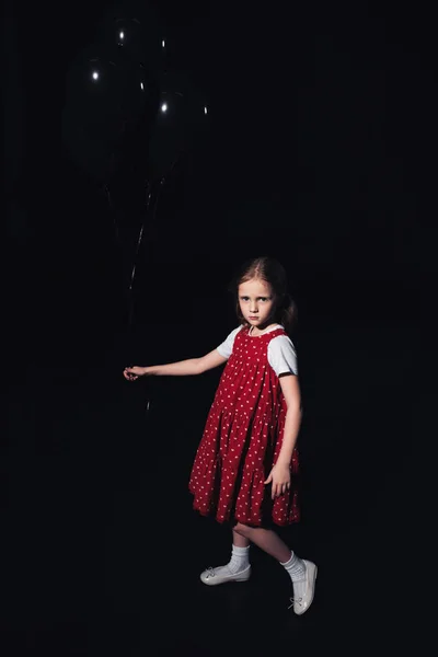 Ofendido niño sosteniendo globos negros y mirando a la cámara aislado en negro - foto de stock