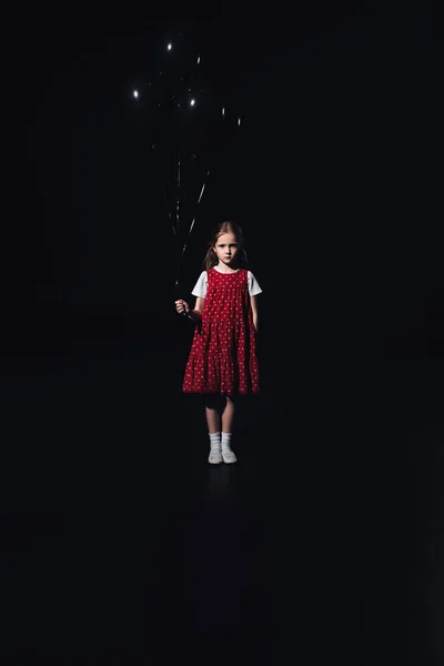 Sad child standing with black balloons and looking at camera isolated on black — Stock Photo