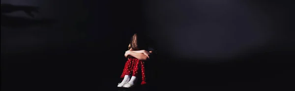 Panoramic shot of depressed child sitting with bowed head on black background — Stock Photo