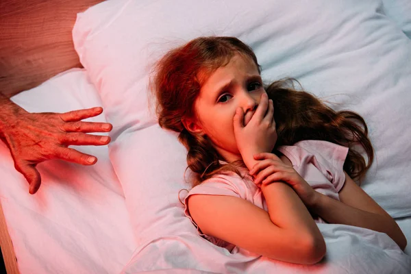 Mano maschile vicino bambino spaventato mostrando segno di silenzio e guardando la fotocamera mentre sdraiato a letto — Foto stock
