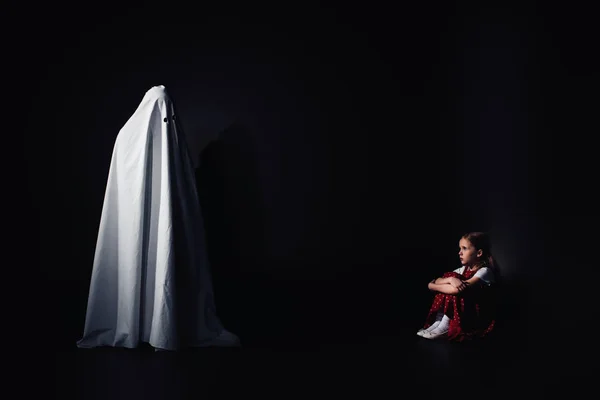 Scared child sitting on floor and looking at terrible white ghost on black background — Stock Photo