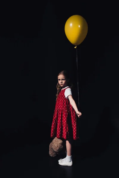 Criança ofendida segurando balão amarelo e ursinho de pelúcia enquanto olha para a câmera isolada no preto — Fotografia de Stock