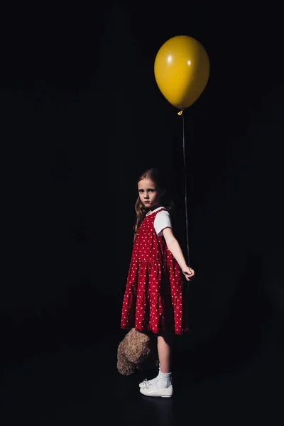 Offensé enfant tenant ballon jaune et ours en peluche tout en regardant la caméra isolée sur noir — Photo de stock