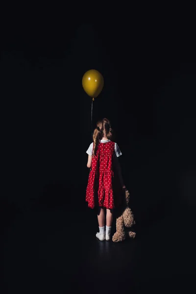 Back view of lonely child with yellow balloon and teddy bear isolated on black — Stock Photo