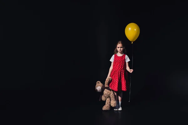 Trauriges Kind mit gelbem Ballon und Teddybär, das isoliert auf schwarz in die Kamera blickt — Stockfoto