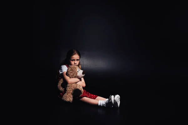 Solitario, niño asustado sentado en el suelo y abrazando osito de peluche sobre fondo negro - foto de stock