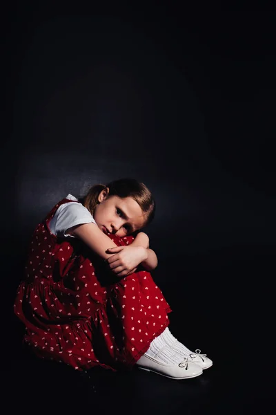 Lonely, frightened child sitting on floor and looking at camera on black background — Stock Photo