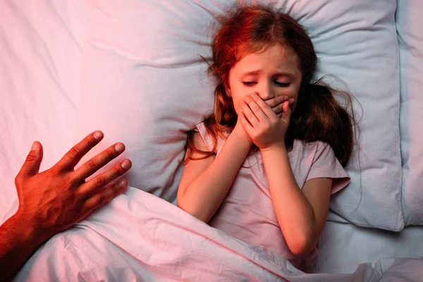 Mano masculina cerca de niño asustado acostado en la cama y mostrando signo de silencio - foto de stock