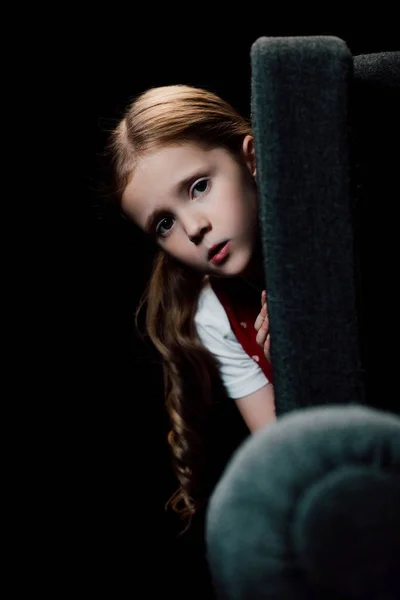 Besorgtes Kind blickt in die Kamera, während es sich isoliert hinter einem schwarzen Sessel versteckt — Stockfoto
