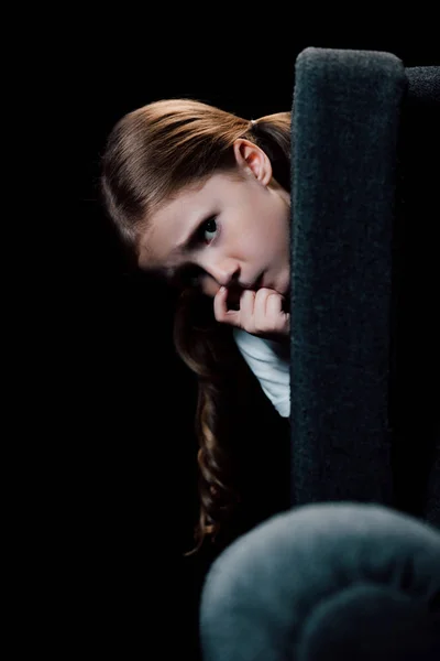 Criança assustada olhando para a câmera enquanto se esconde atrás de poltrona isolada no preto — Fotografia de Stock