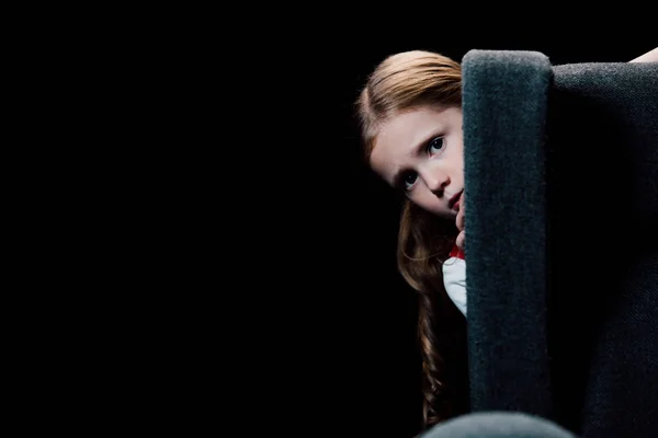 Scared child looking at camera while hiding behind armchair isolated on black — Stock Photo