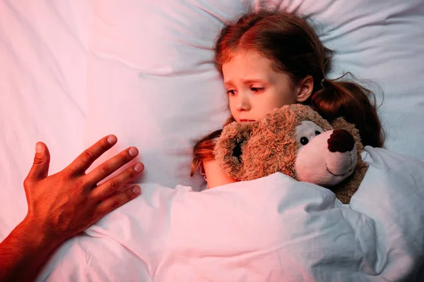 Mano masculina cerca de niño asustado acostado en la cama con oso de peluche - foto de stock