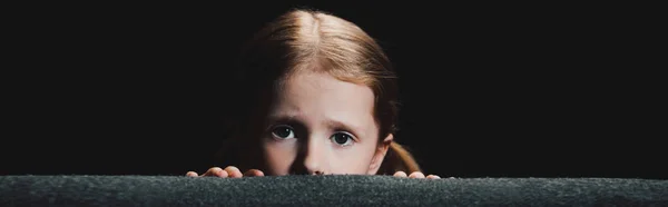 Panoramic shot of frightened child hiding behind armchair and looking at camera  isolated on black — Stock Photo