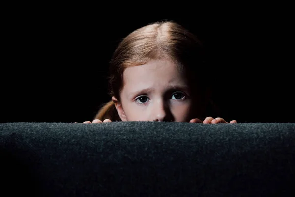 Niño asustado escondido detrás del sillón y mirando a la cámara aislada en negro - foto de stock