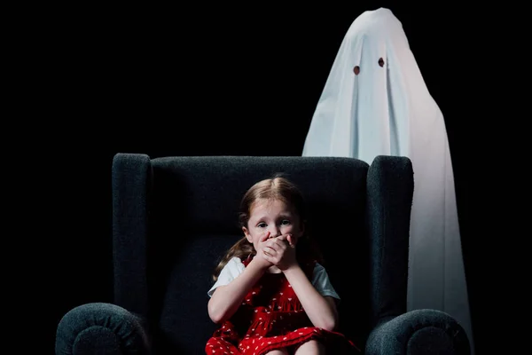 White ghost and scared child sitting in armchair and showing hish sign isolated on black — Stock Photo