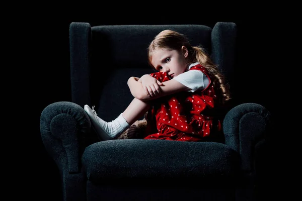 Niño asustado y solitario sentado en un sillón y mirando a la cámara aislada en negro - foto de stock