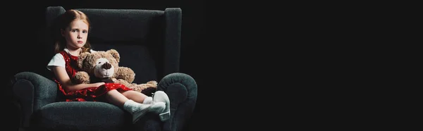 Triste, solitaire enfant assis dans un fauteuil avec ours en peluche et regardant caméra isolé sur noir — Photo de stock