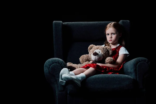 Trauriges, einsames Kind im Sessel sitzend und Teddybär isoliert auf schwarz haltend — Stockfoto