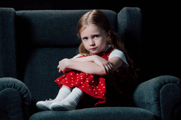 Enfant déprimé et effrayé assis dans un fauteuil et regardant la caméra isolée sur noir — Photo de stock