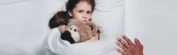 Partial view of male hand near scared child lying in bed with teddy bear, panoramic shot — Stock Photo