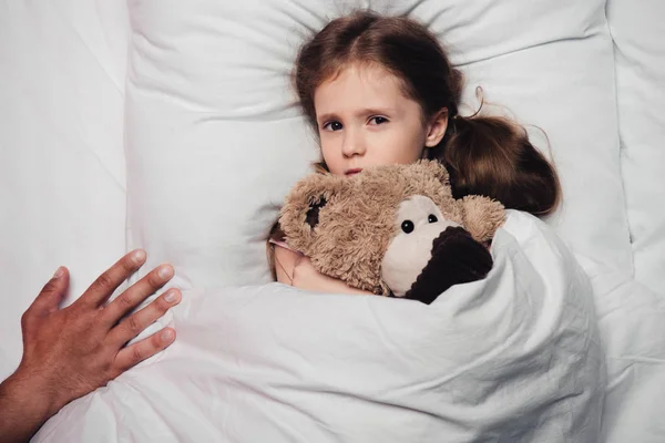 Обрезанный вид мужской руки рядом с испуганным ребенком, лежащим в постели с плюшевым мишкой — стоковое фото