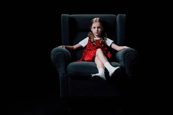 Самотня дитина сидить у кріслі і дивиться на камеру ізольовано на чорному — стокове фото