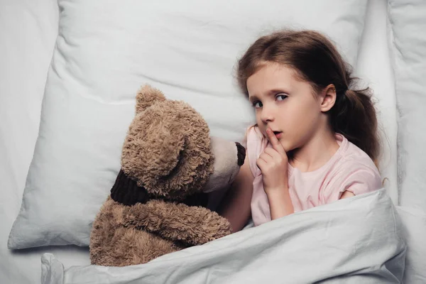 Vue de dessus de l'enfant effrayé montrant le signe de silence et regardant la caméra tout en étant couché dans le lit avec l'ours en peluche — Photo de stock