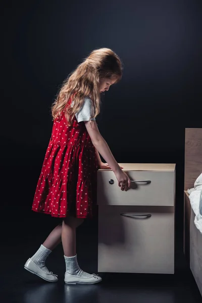 Cute child in red dress opening nightstand on black background — Stock Photo