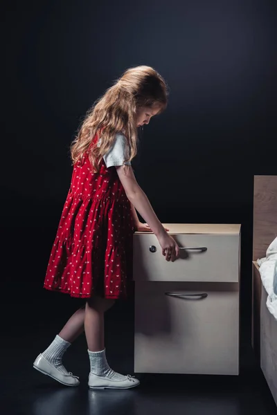 Niedliches Kind im roten Kleid öffnet Nachttisch auf schwarzem Hintergrund — Stockfoto