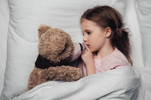 Adorable enfant montrant signe de silence tout en étant couché dans le lit avec ours en peluche — Photo de stock