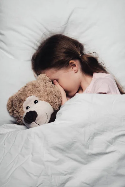 Очаровательный ребенок закрывает глаза рукой, лежа в постели с плюшевым мишкой — стоковое фото
